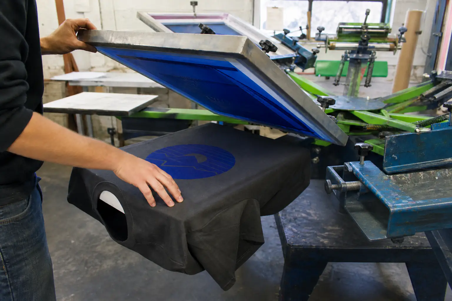 Print operator lifting a screen to inspect a cobalt print on a manual carousel